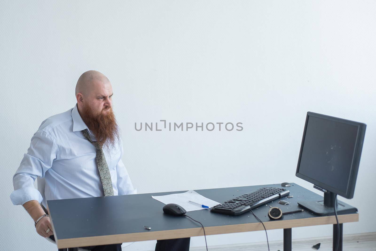 Angry bald man with a red beard in the office in a business suit crashes a computer. Manager with a nervous breakdown breaks the keyboard on the monitor