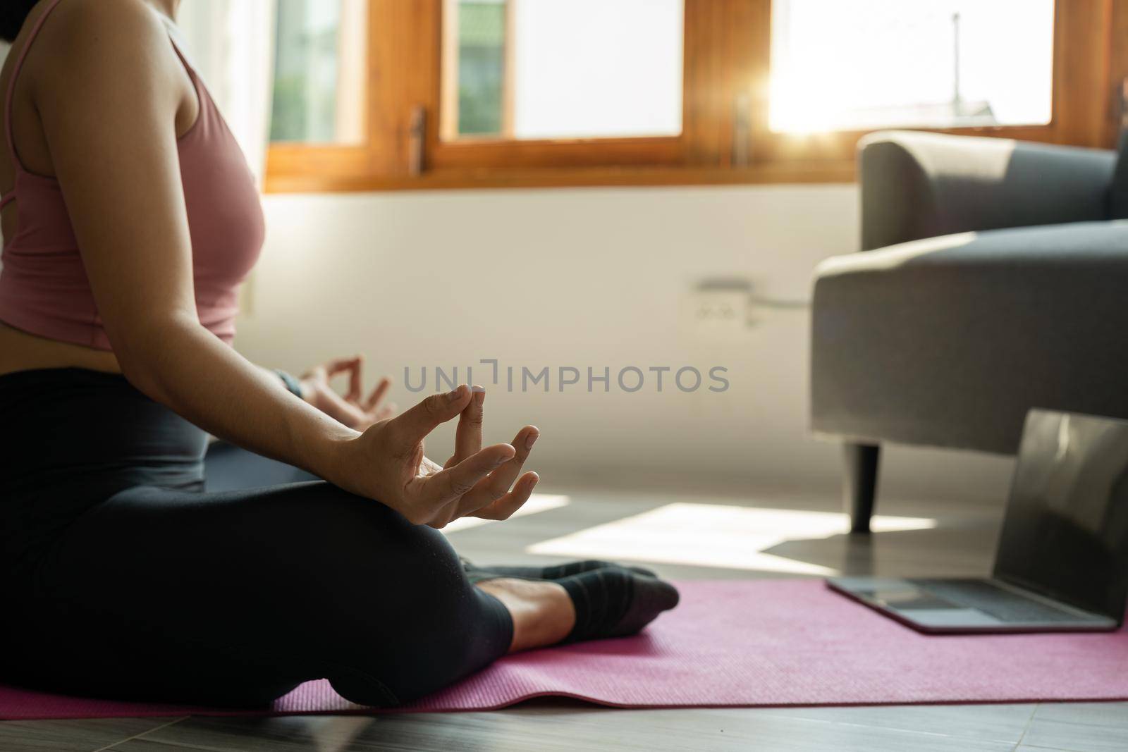 Asian woman meditating with trainer online via laptop connection. Healthy lady sitting on the floor on yoga mat in lotus position.