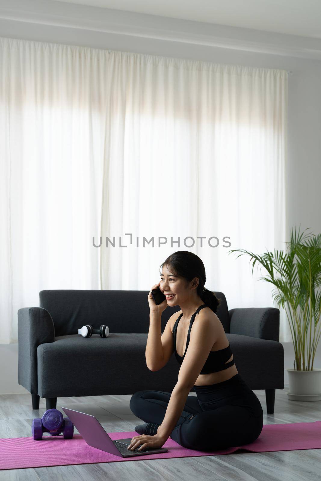 Portrait asian sporty woman in sportswear working out and using laptop and call phone at home in living room, sitting on the floor with dumbbells on yoga mat. Sport and online training concept by nateemee