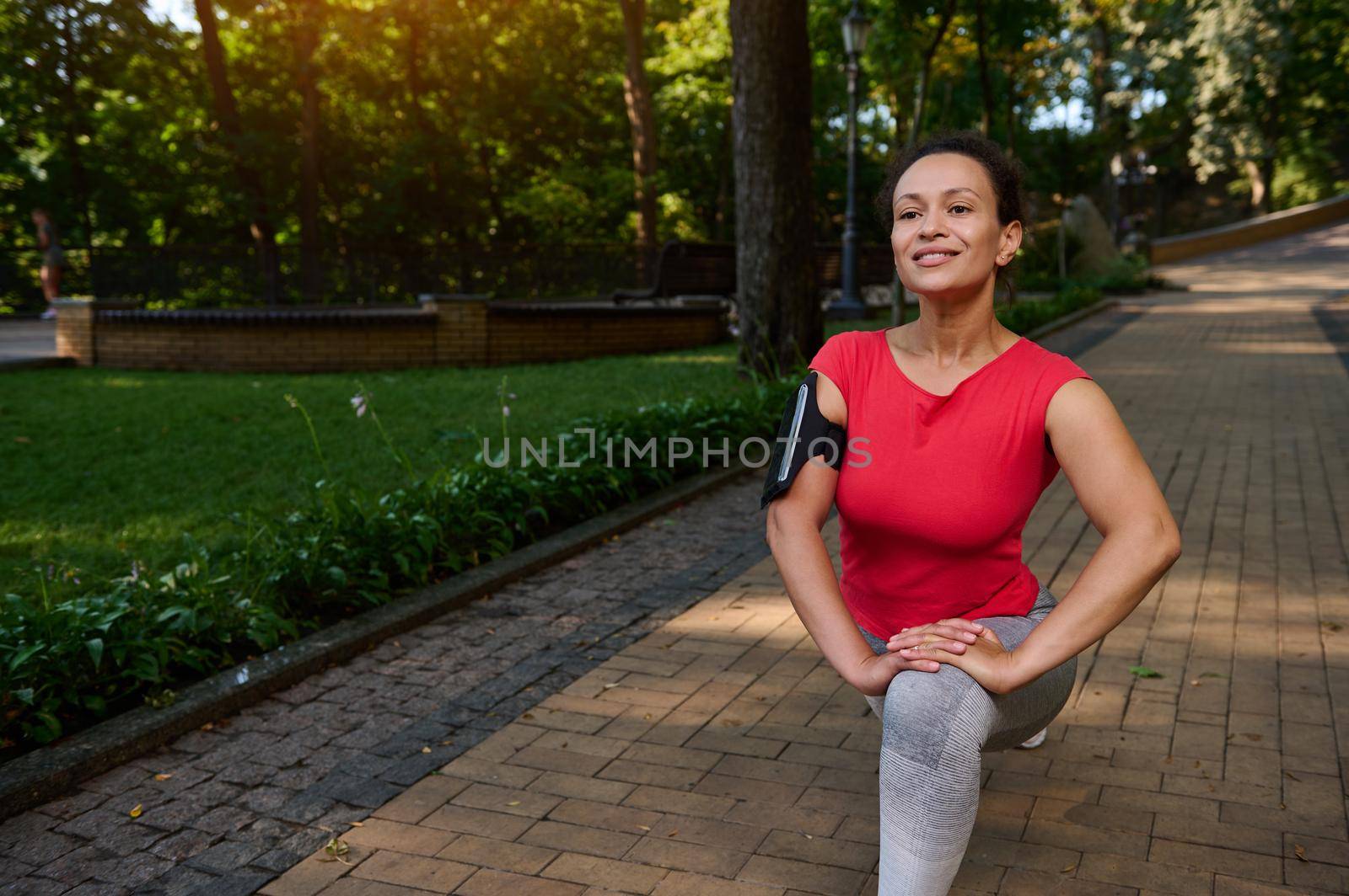 Beautiful middle aged sporting woman exercising outdoor, doing lunges, stretching her leg muscles while exercising in the city park on warm sunny summer day