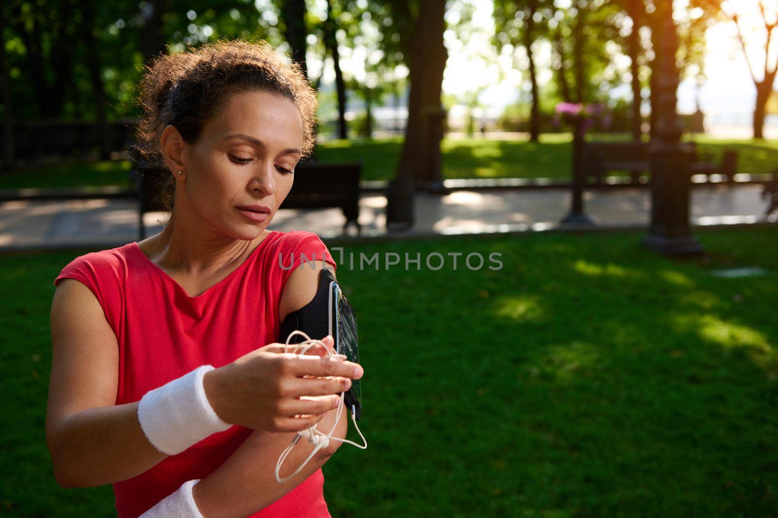 Middle aged beautiful African woman, sportswoman, athlete adjusting the mobile app in her mobile phone in the smartphone holder, standing in the city park, getting ready for morning jog