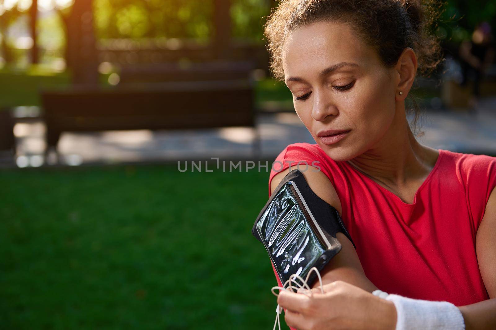 Close-up portrait of a young athletic woman, a runner in a bright red t-shirt, connects headphones to a mobile phone and sets up the application before jogging in the park on a sunny summer day