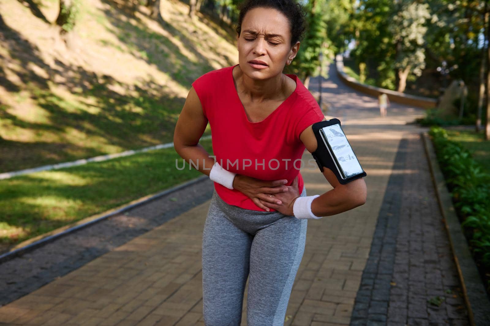Female runner, African American sportswoman feeling exhaustion and side cramps, stomach-ache during running in the city park. Healthy active lifestyle concept, sport injury