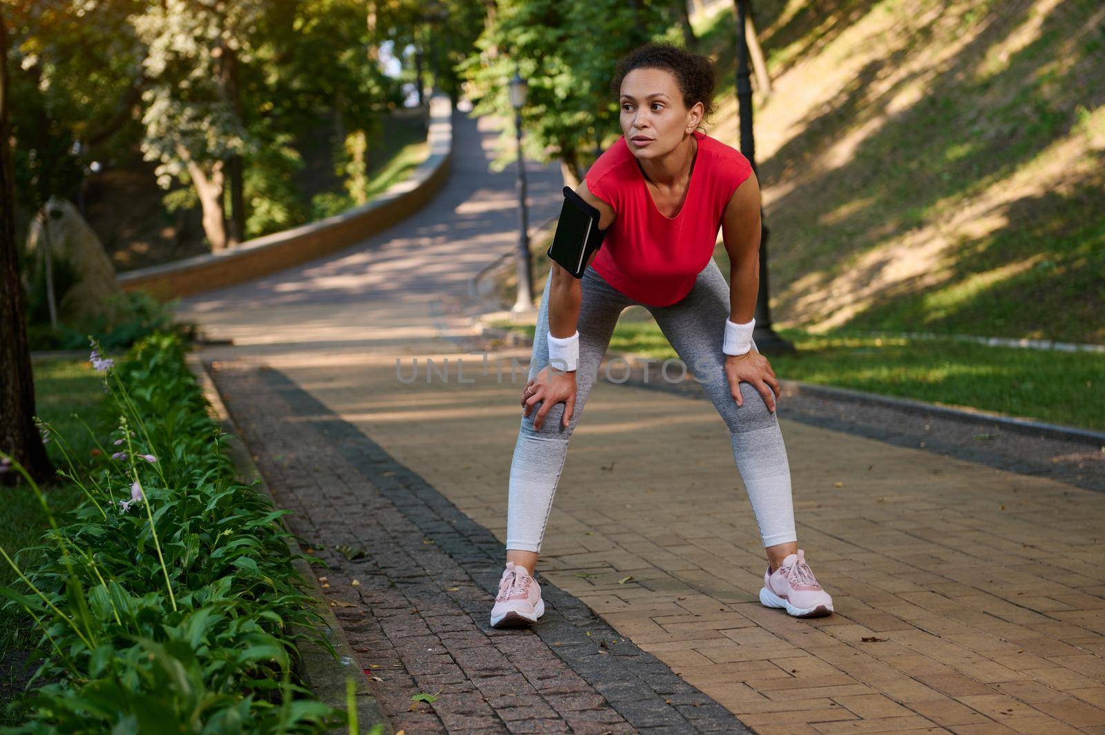 Female runner, athlete, sportswoman resting in the city park while feeling exhausted during jogging on the treadmill on a warm sunny summer day. Body weight training, slimming and dieting concept