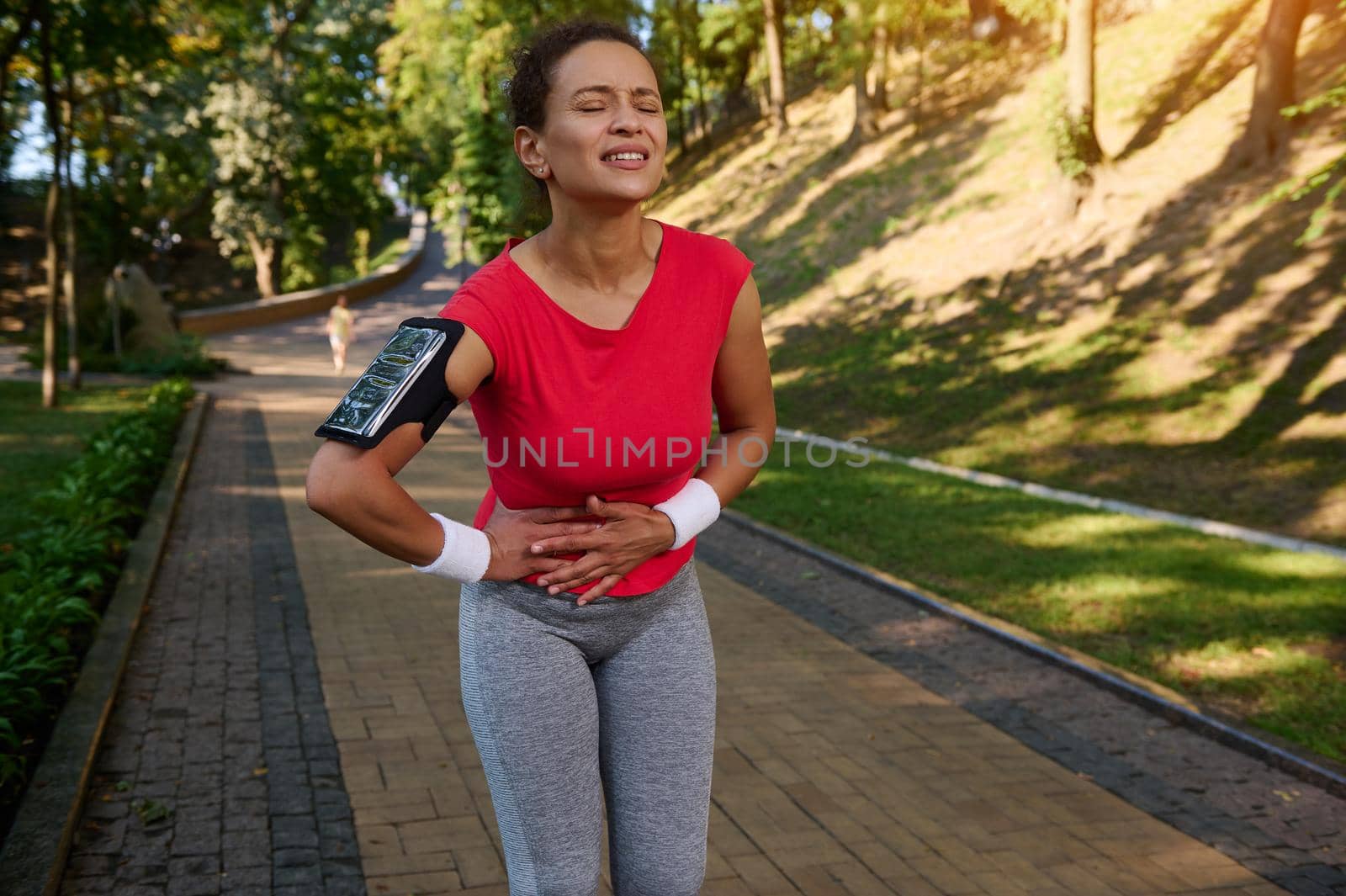 Beautiful female runner athlete in tight sportswear and with a smartphone holder, putting her hands on her stomach, feeling cramps in her side while running on a treadmill in a city park. Sport injury
