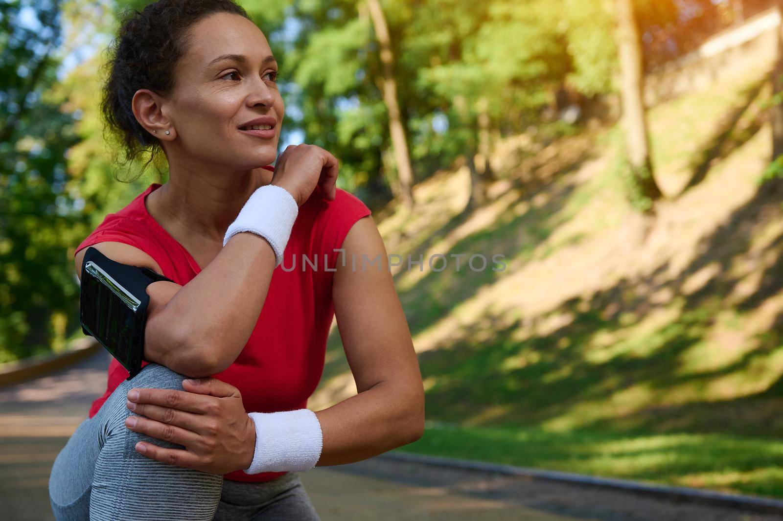 Close-up portrait of beautiful Hispanic woman, athlete in sportswear relaxing during jog in the city park, smiles looking aside, enjoying morning workout on summer sunny day