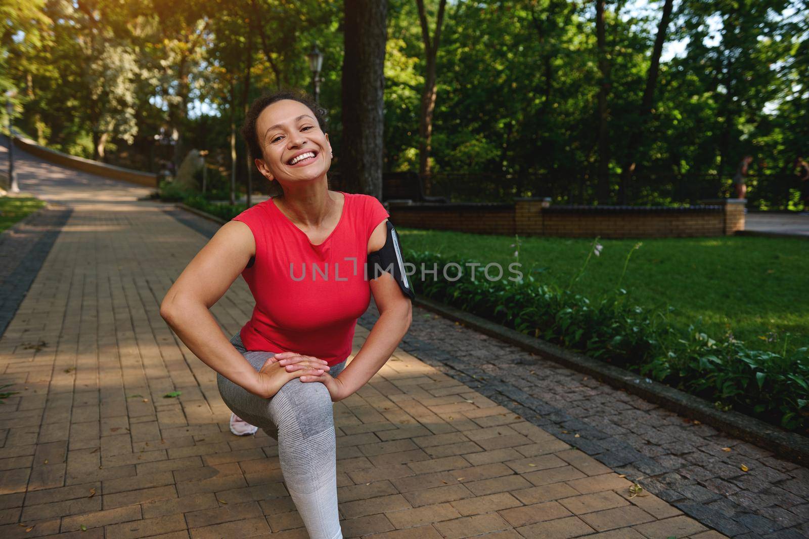 Happy cheerful African woman doing lunges while exercising outdoor, smiles cutely with beautiful toothy smile looking at camera, enjoying her active and healthy lifestyle