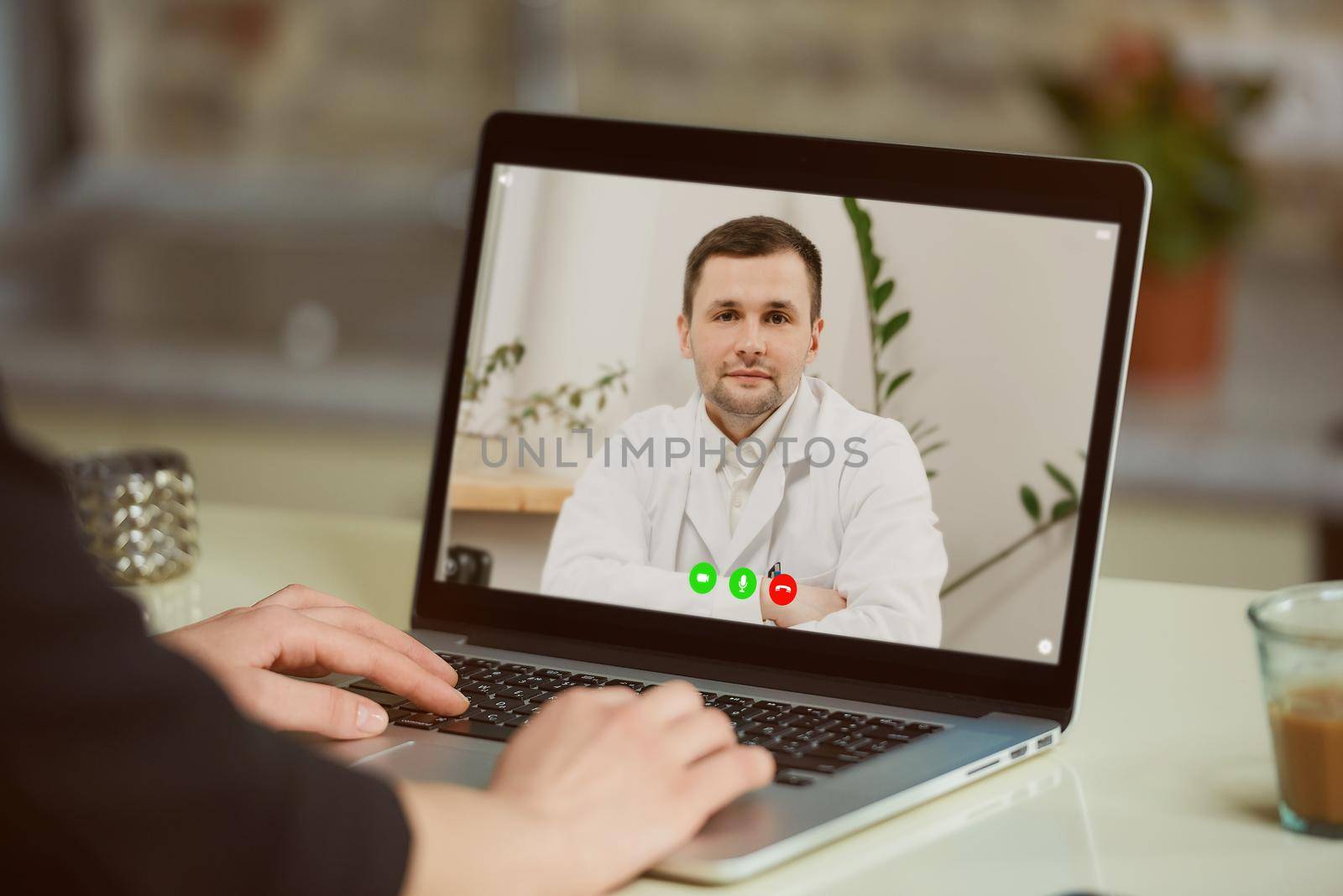 A laptop screen view over a woman's shoulder. A woman at an online doctor's appointment. A girl is discussing her health with a therapist in a white lab coat on a video call.