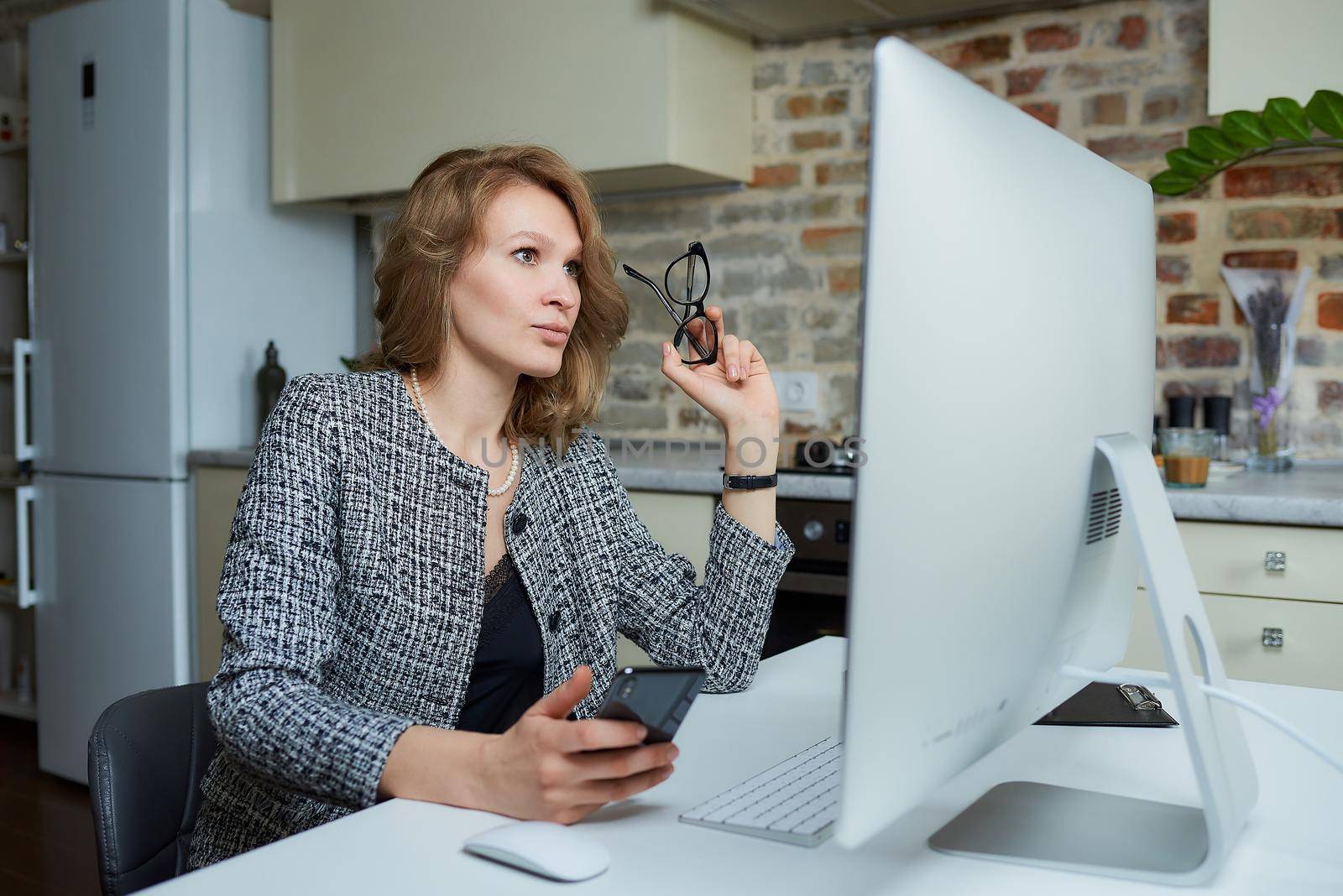 A woman with glasses works remotely on a desktop computer in her studio. A lady uses a phone during a video conference at home. A female professor listening to student's answers on an online lesson.