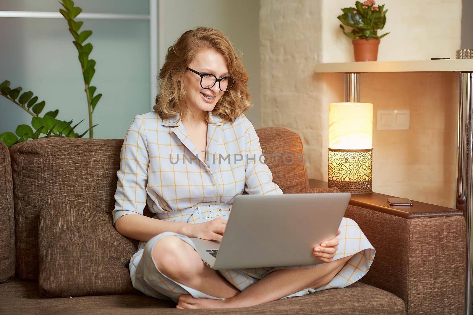 A young woman in a dress sitting on the couch with legs crossed works remotely on a laptop in her apartment. A female student with braces in glasses laughing and listening to an online lecture.