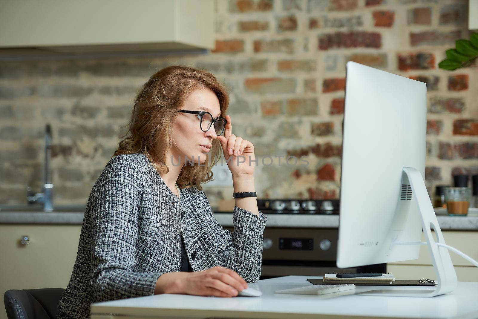 A woman in glasses works remotely on a desktop computer in her studio. A female boss searching for information at a video conference at home. A pretty female professor preparing for an online lecture.