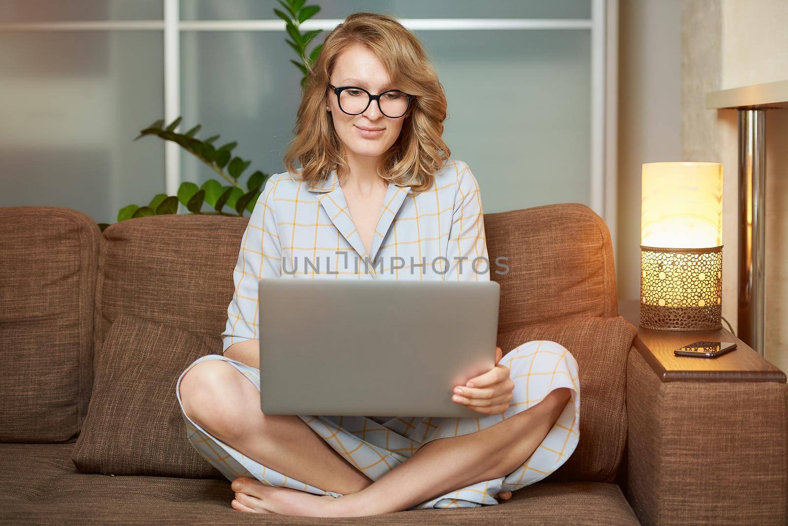 A young woman in glasses works remotely on a laptop in her apartment. A cute girl during a video conference with her colleagues at home. A female student with a smile listening to an online lecture.