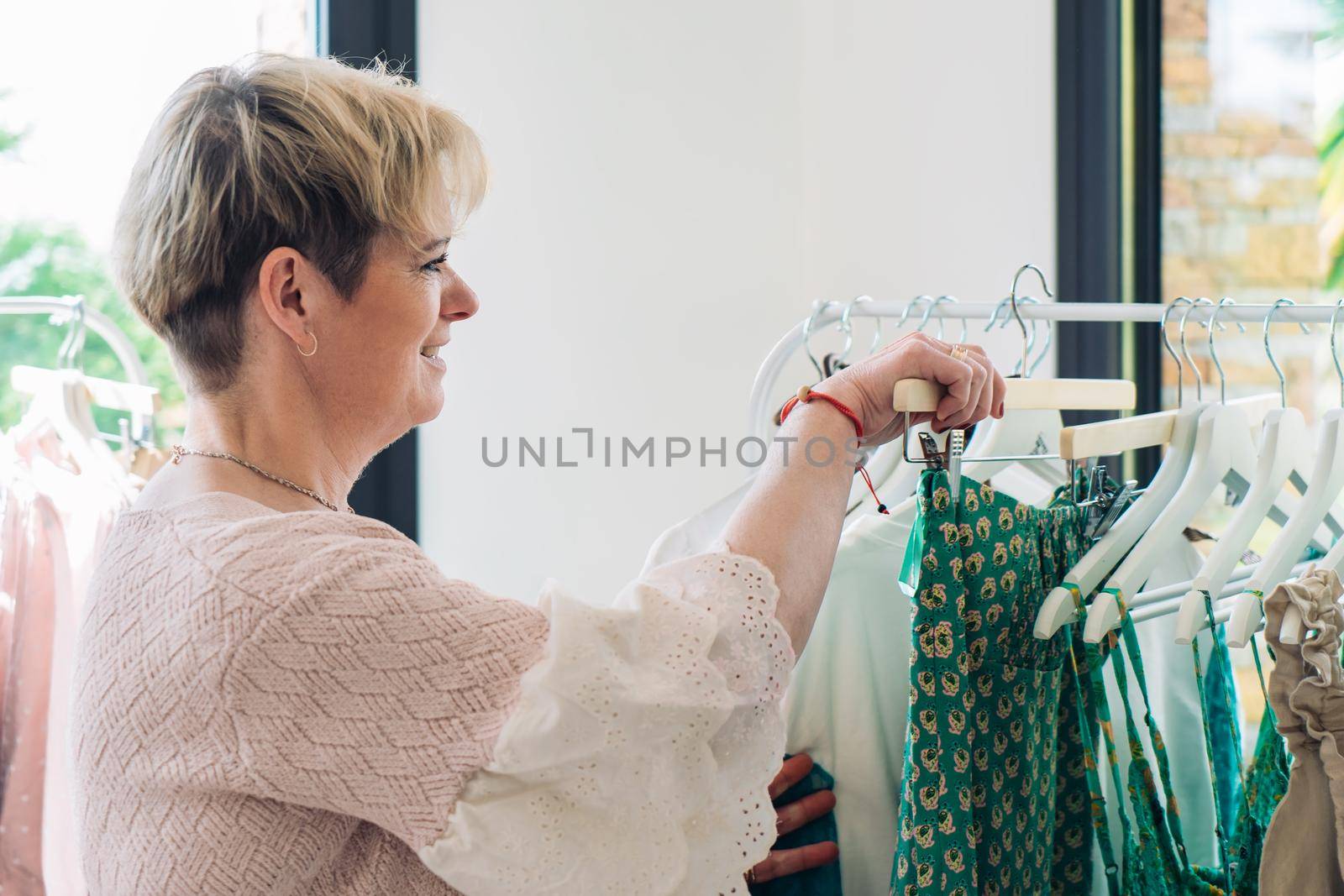 blonde mature woman happy and enjoying a day of shopping, shopping for clothes in a fashion shop. shopping concept natural light from the shop window, sun rays, display with clothes, rack, shop window, clothes, Horizontal.