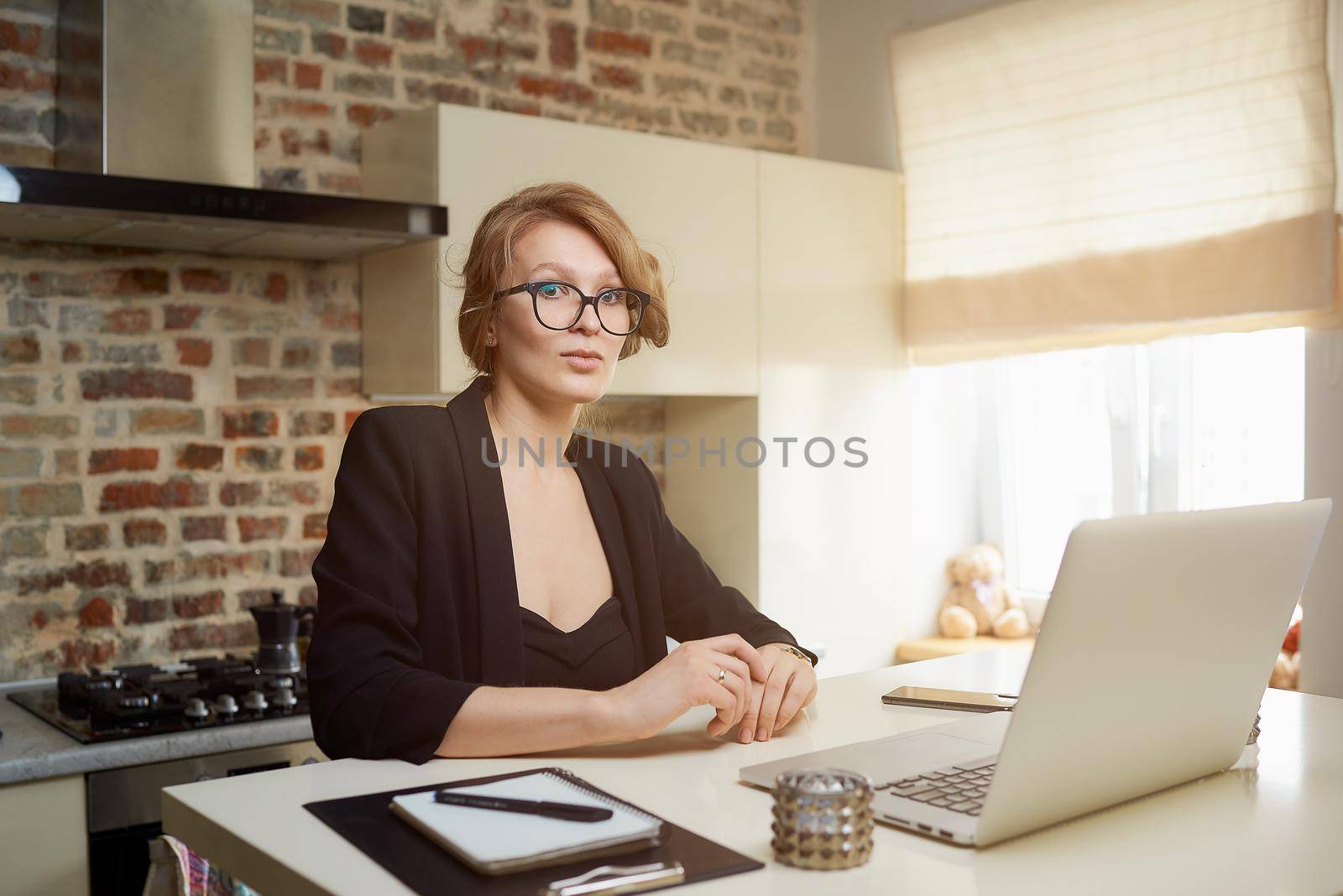 A young woman in glasses works remotely on a laptop in her kitchen. A blond girl listening to her colleagues on a video conference at home. A lady studying a lesson online on a webinar.