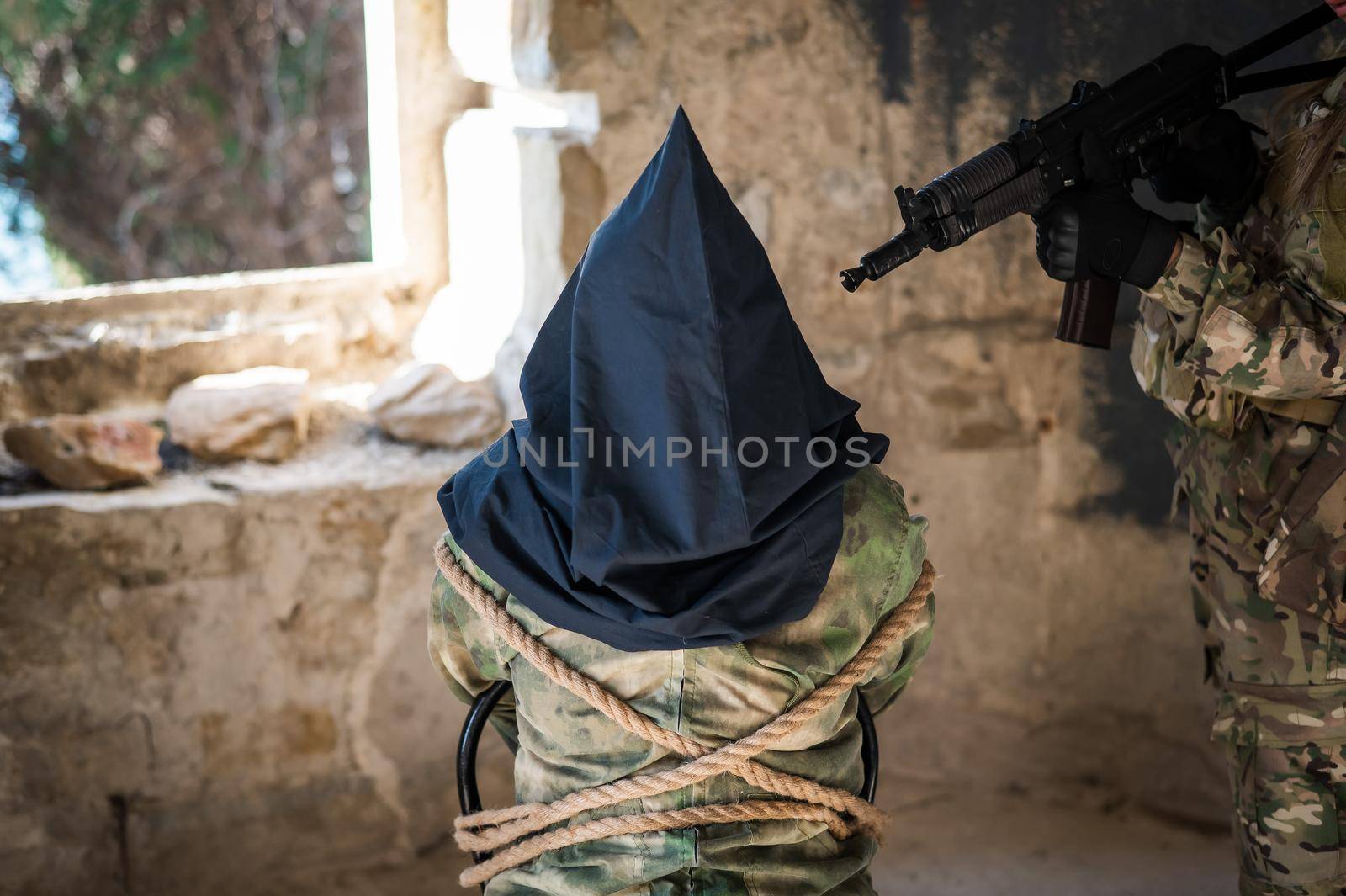A caucasian woman in an army uniform holds a hostage with a bag on her head at the sight of a machine gun