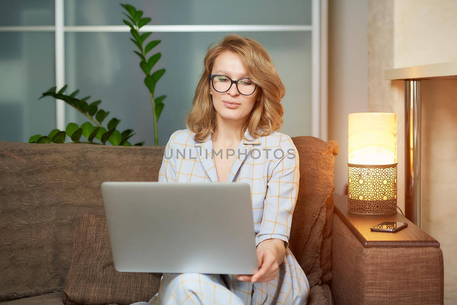 A young woman in glasses works remotely on a laptop in her apartment. A lady during a video business briefing at home. A pretty female student listening to an online lecture.