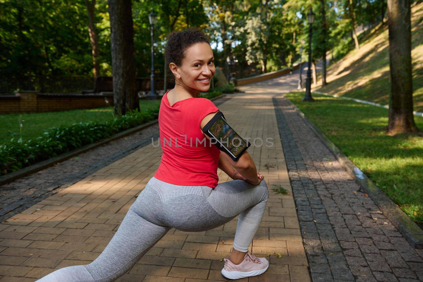 View from the back to an African woman doing lunges while exercising outdoor, smiles cutely with beautiful toothy smile looking at camera, enjoying her active and healthy lifestyle