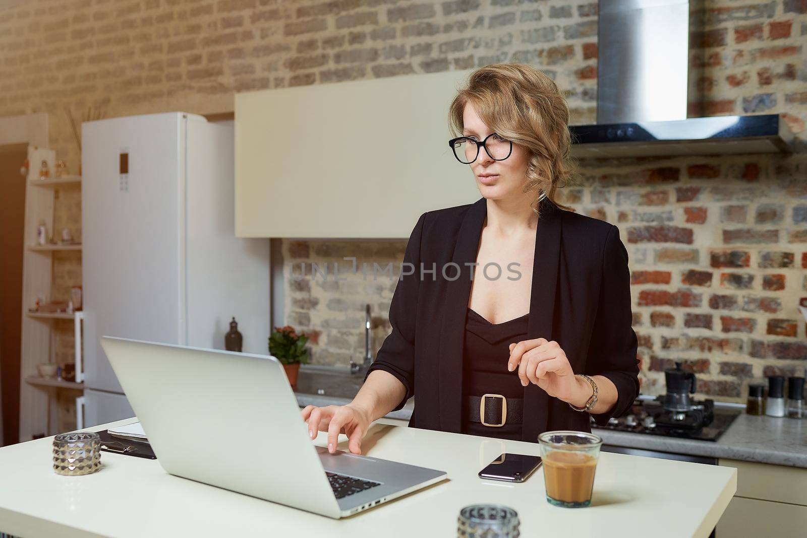 A young woman in glasses works remotely on a laptop in her kitchen. A serious girl calmly browsing news on the internet at home. A lady preparing for a lecture on a video call..