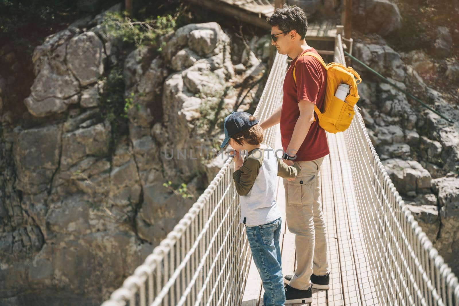 Father and son looking afar from the rope bridge in the mountains when hiking. Casually dressed tourists child school boy and his dad with yellow backpack crossing the canyon through rope bridge.