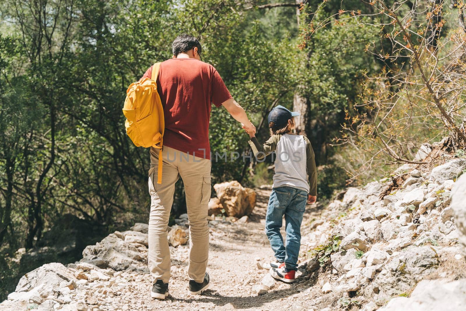 Rear view of tourists school boy and his dad walking a stone footpath in spring forest. Child kid and father wearing casual clothes and yellow backpack while hiking in summer greenwood leaf forest by Ostanina