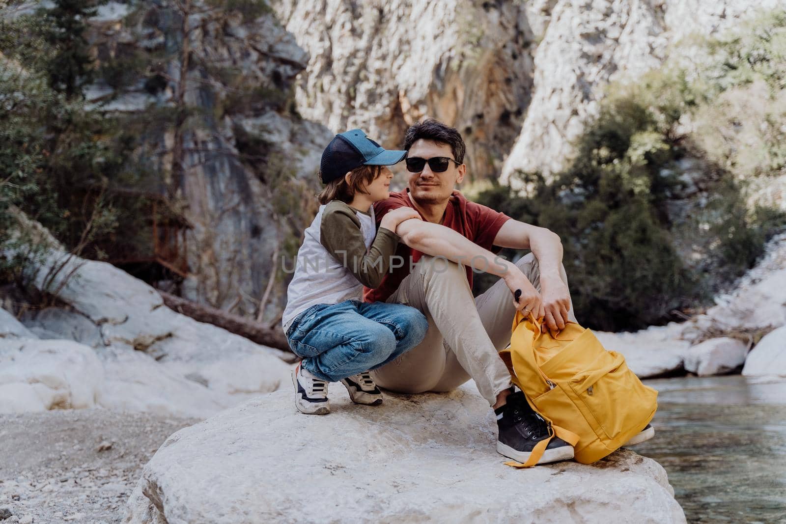 School boy and his dad with yellow backpack sits on a riverside rock in the canyon with mountain cliffs in the background. Kid child and father taking a rest on a boulder near mountain river.