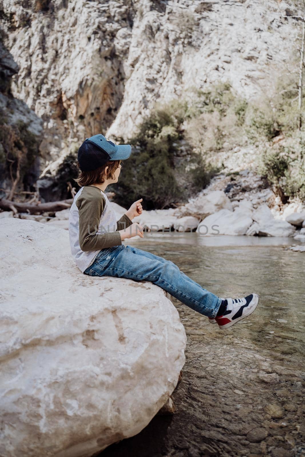 Caucasian school boy sits on a riverside rock in the canyon with mountain cliffs in the background. Kid child taking a rest on a boulder near mountain river bank by Ostanina