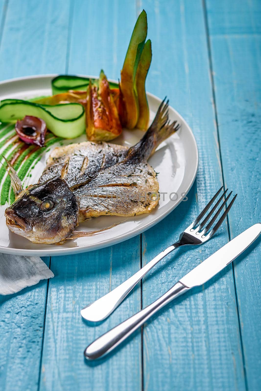 Fried fish dorado, decorated with cucumbers and onions. The original presentation of the dish from the chef. On a blue wooden background