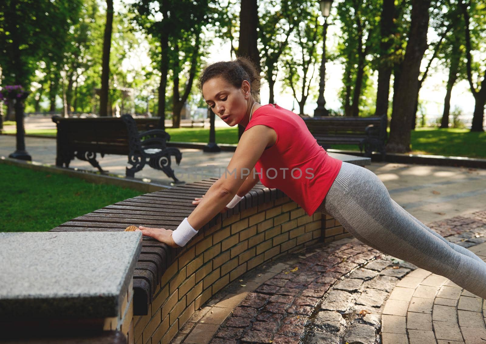 Determined motivated middle aged muscular Hispanic woman, sportswoman exercising outdoors, doing push ups, enjoying morning functional cross workout in city park on a beautiful hot sunny summer day