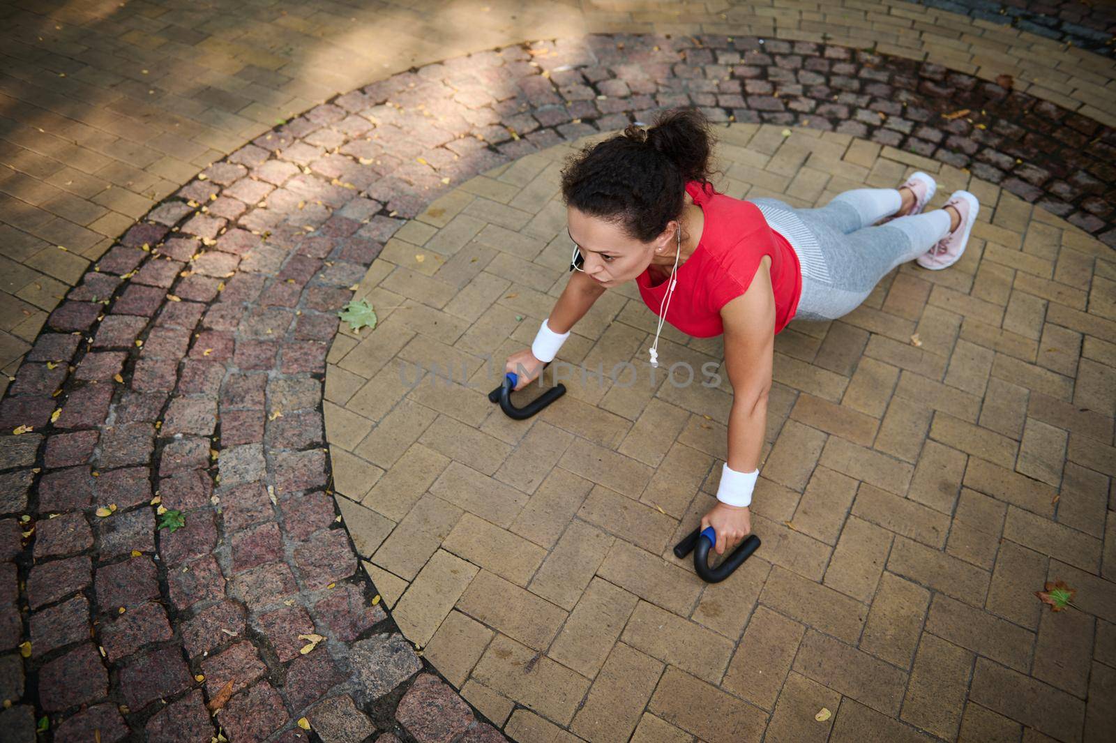 Determined and motivated middle aged African woman of average build in tight sportswear doing push-ups from the floor during a morning workout in a city park on a warm summer day