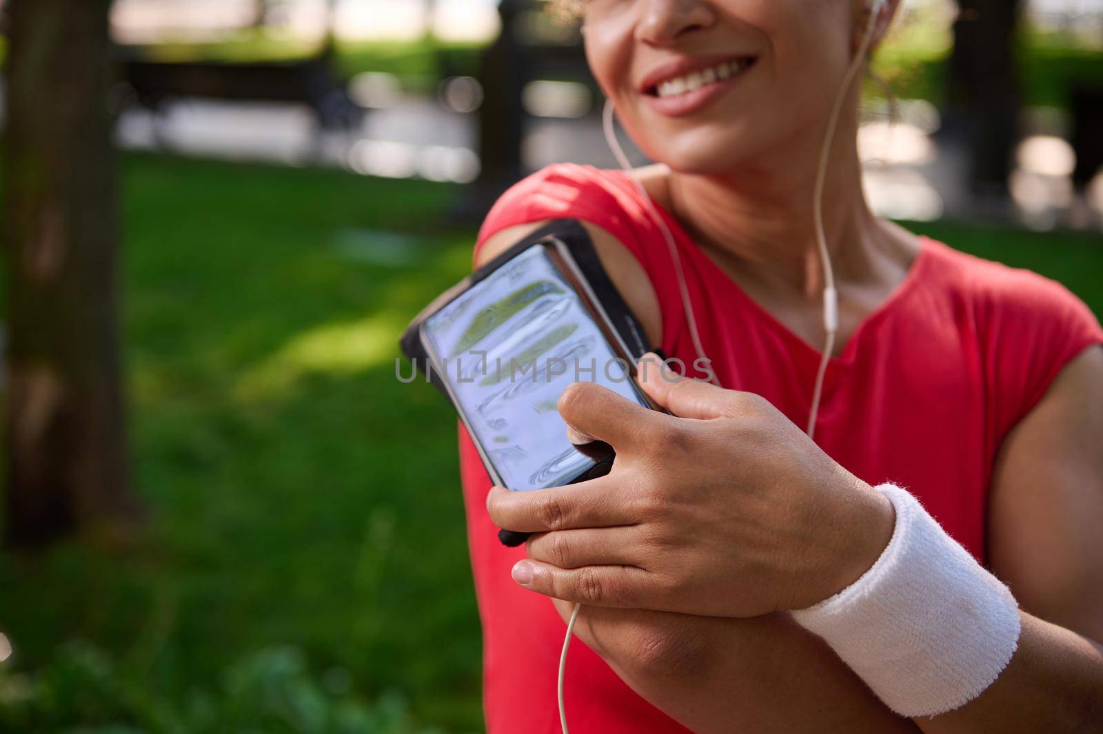 Soft focus on a smartphone holder on the arm of blurred charming smiling female athlete, putting her finger on the phone and adjusting mobile app and playlist during morning jog. Sport and gadgets