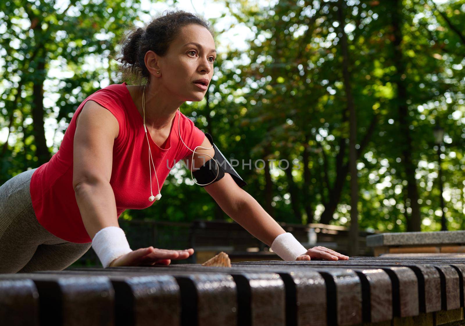 Close-up of a determined Hispanic female athlete, muscular build woman in bright sportswear performing push ups from wooden bench in the city park during functional cross training in the morning