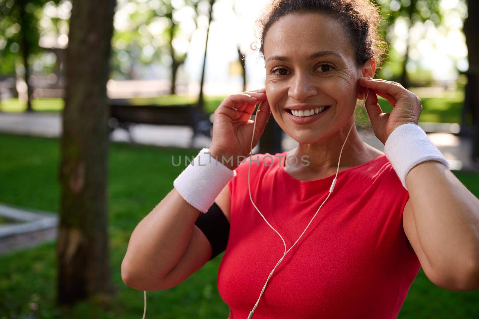 Charming African American sportswoman, determined athlete, runner in bright red t-shirt and white terry wristbands smiles to camera while putting on earphones before morning run on sunny summer day