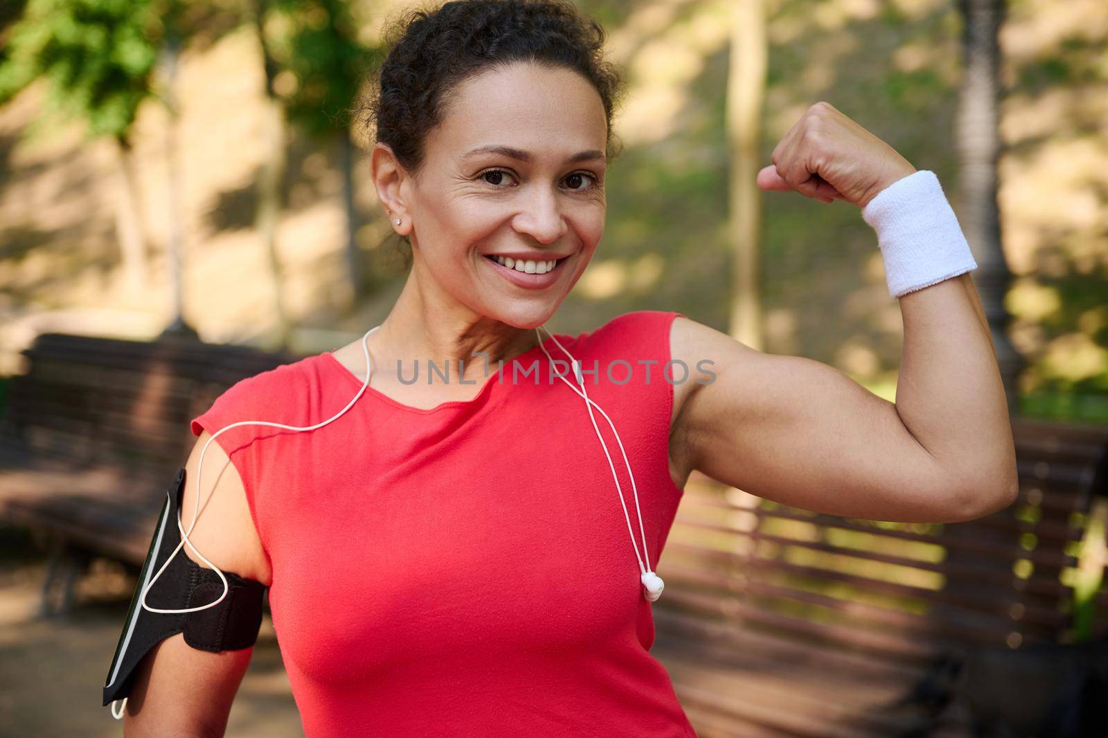 Go beyond your limits. Active multi ethnic middle aged athlete muscular sportswoman smiling beautiful toothy smile and showing her toned biceps to camera during workout. Sport and healthy body concept