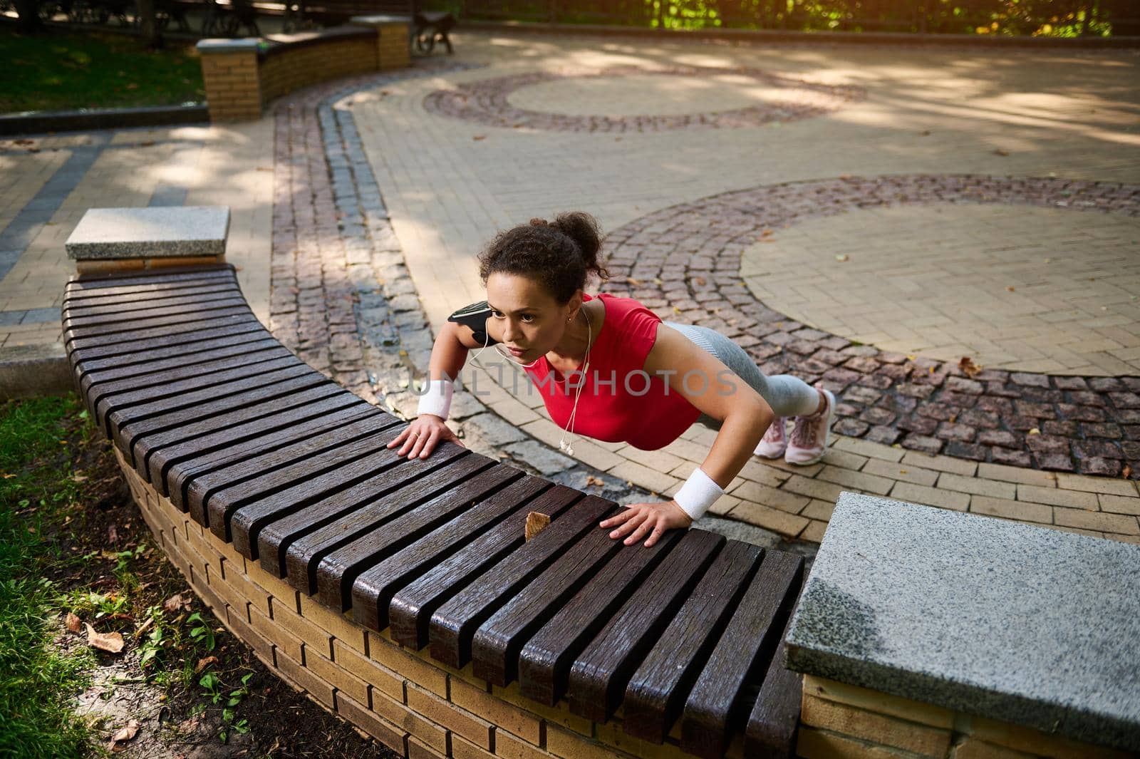 High angle view of a middle-aged African muscular athletic woman, sportswoman exercising outdoors, pushing up from bench during functional cross training in landscaped city park
