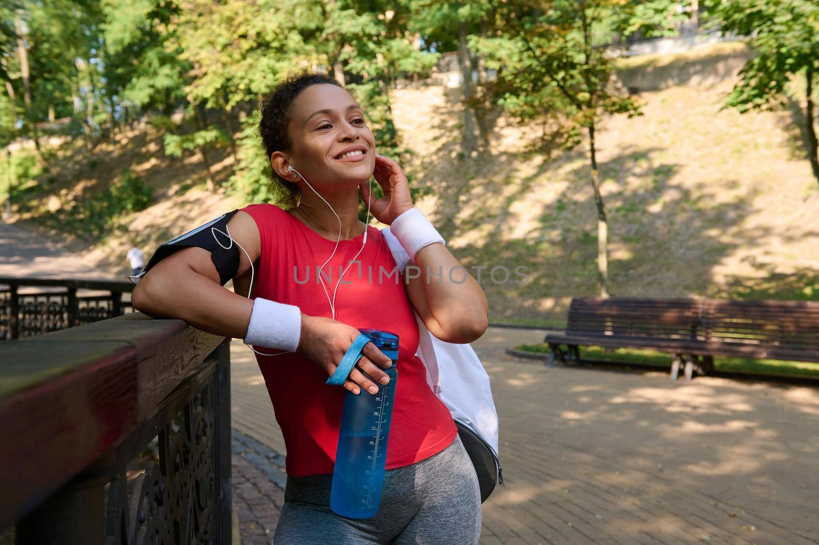 Charming active sportswoman in activewear smiles beautiful toothy smile, holding a bottle with water and putting on earphones, relaxing after morning jog, functional and cardio workout in city park