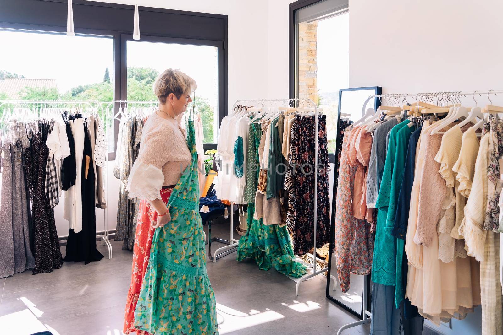 mature woman testing a dress in front of a mirror. blonde female shopping in a fashion shop. shopping concept. leisure concept. Natural light, sunbeams, display, clothes rack, clothes, horizontal view, space to copy.
