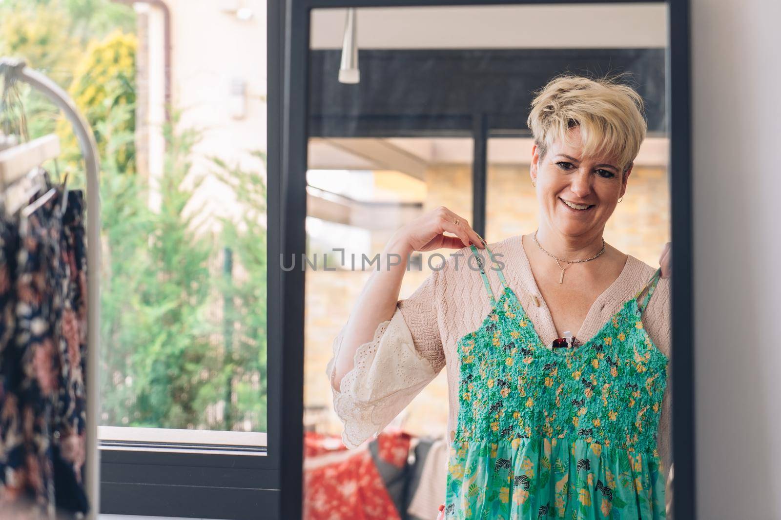 mature woman testing a dress in front of a mirror. blonde female shopping in a fashion shop. shopping concept. leisure concept. Natural light, sunbeams, display, clothes rack, clothes, vertical view, space to copy.