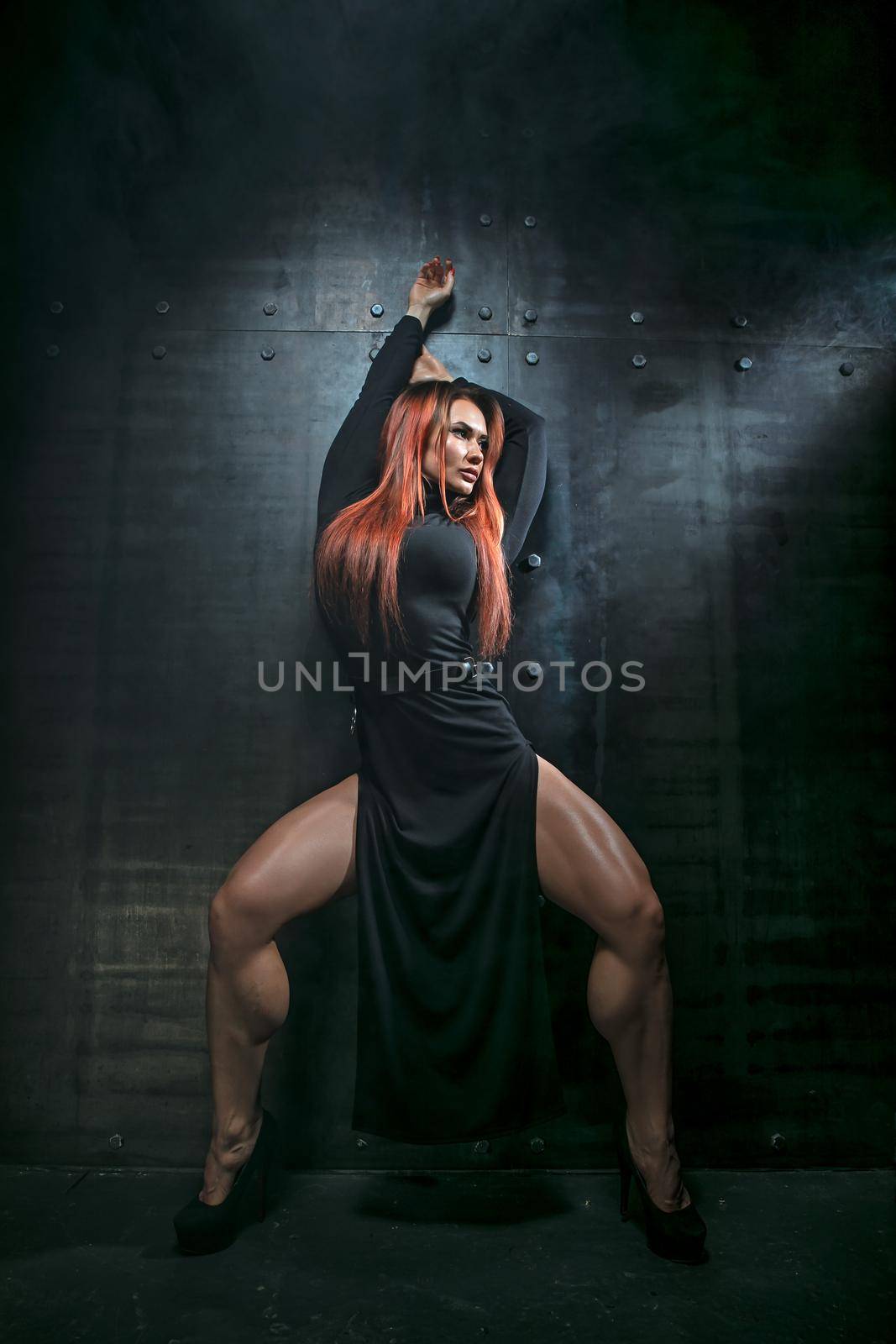 Graceful red-haired girl in black dress with high heels in the studio on metallic background