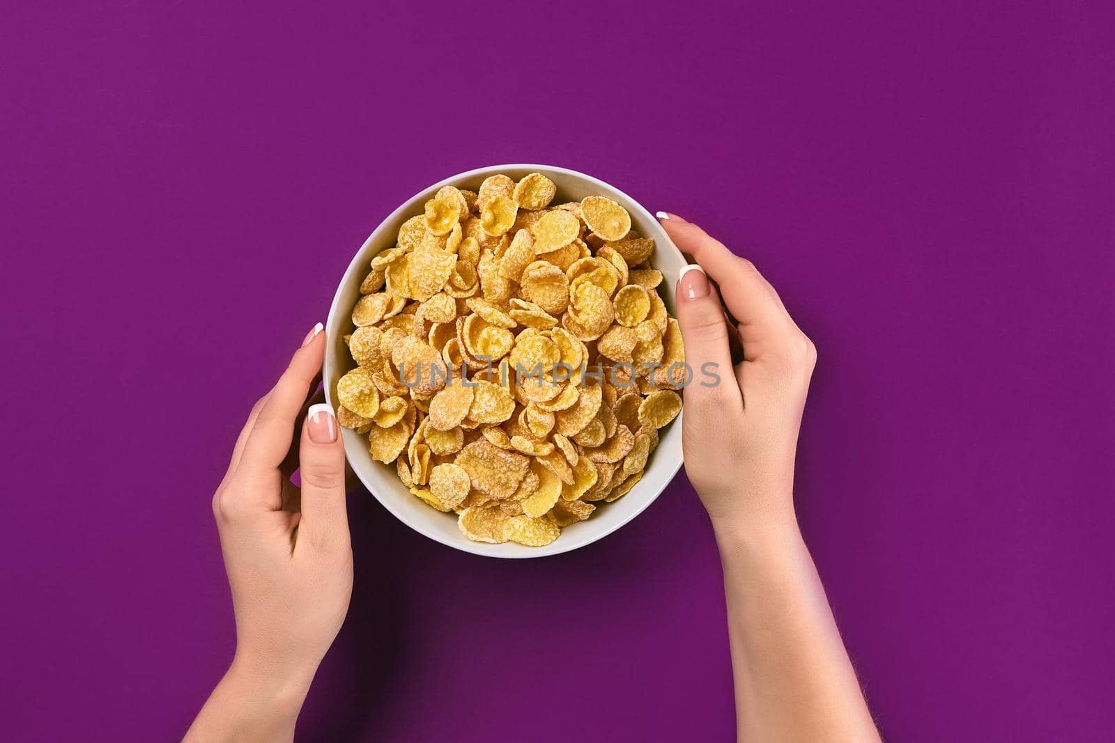 Female hands holding bowl with healthy breakfast, closeup. Bowl with cornflakes on the colorful background. Purple background, top view. Copy space. Still life. Flat lay