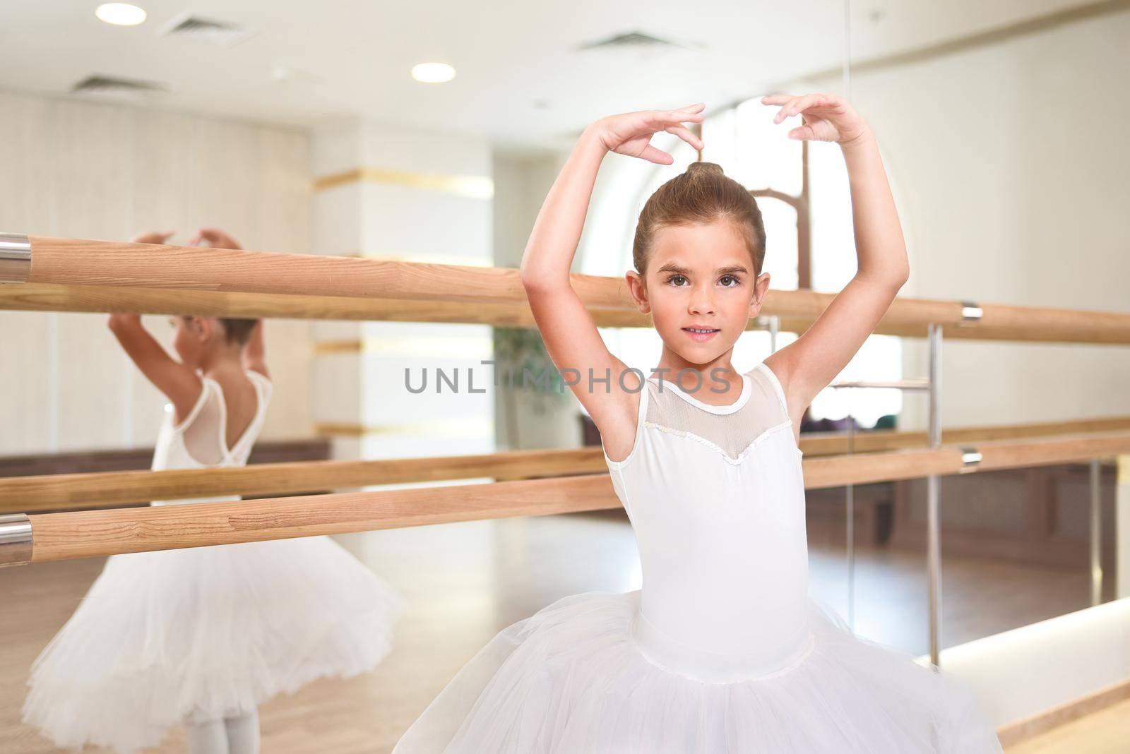 cute little ballerina standing with her hands up in a performance dress. She is dreaming to become a professional ballet dancer. Dance school, ballet studio. by Nickstock