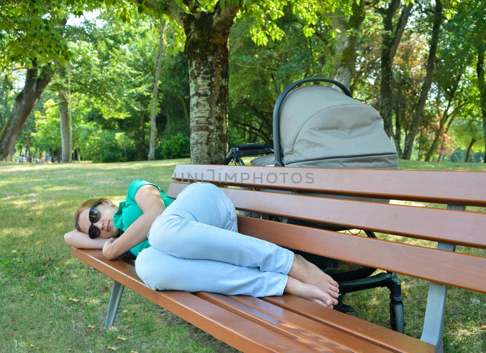 Tired mother asleep on a bench in a park with a baby in a stroller