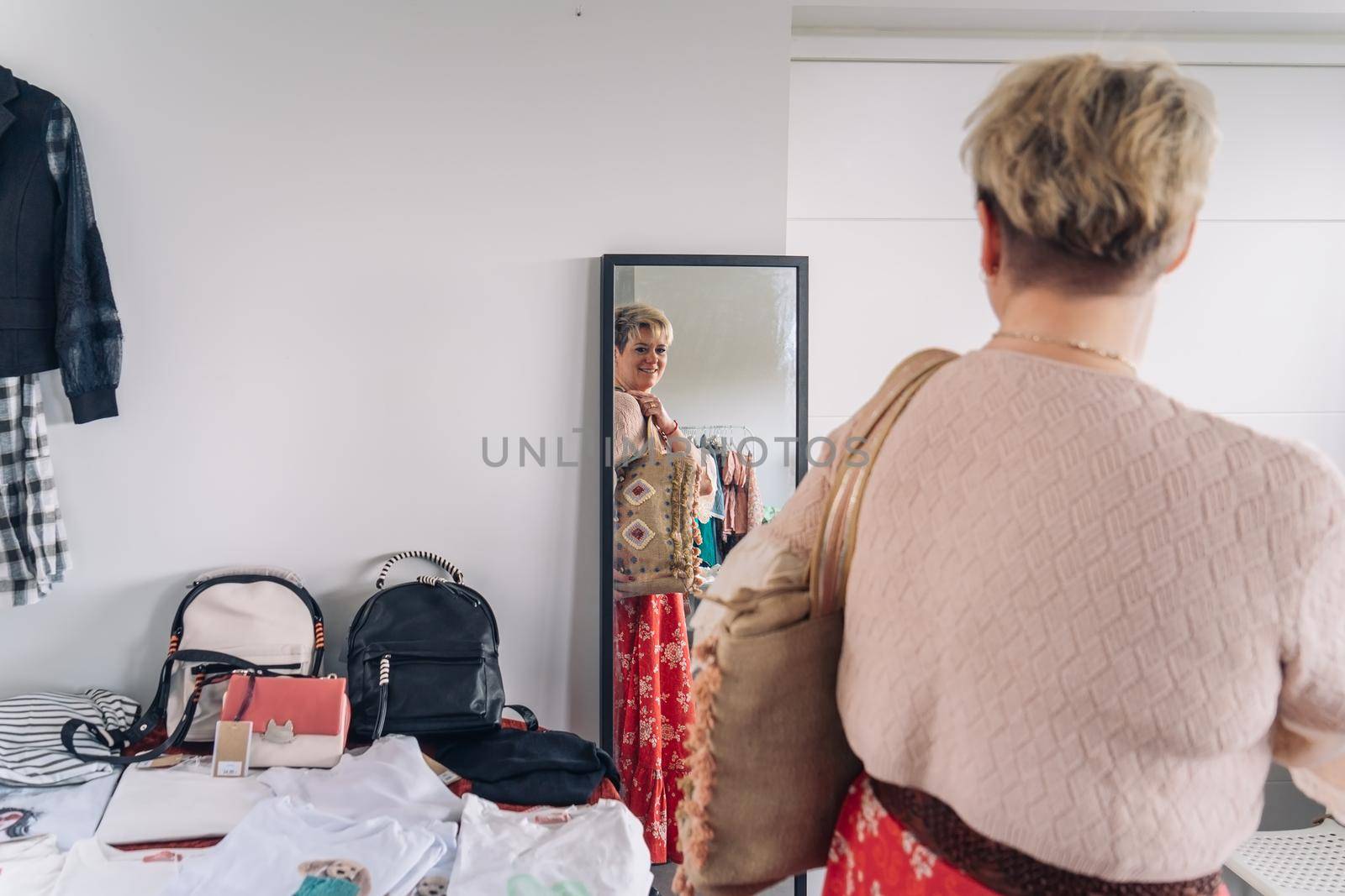 mature woman trying on handbags in front of mirror in fashion shop. blonde woman buying a large handbag. by CatPhotography