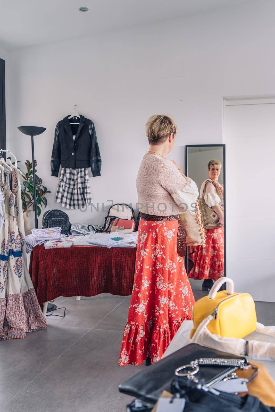 mature woman trying on handbags in front of mirror in fashion shop. blonde woman buying a large handbag. natural light, vertical view, interior, clothes, hangers. concept of shopping. concept of leisure