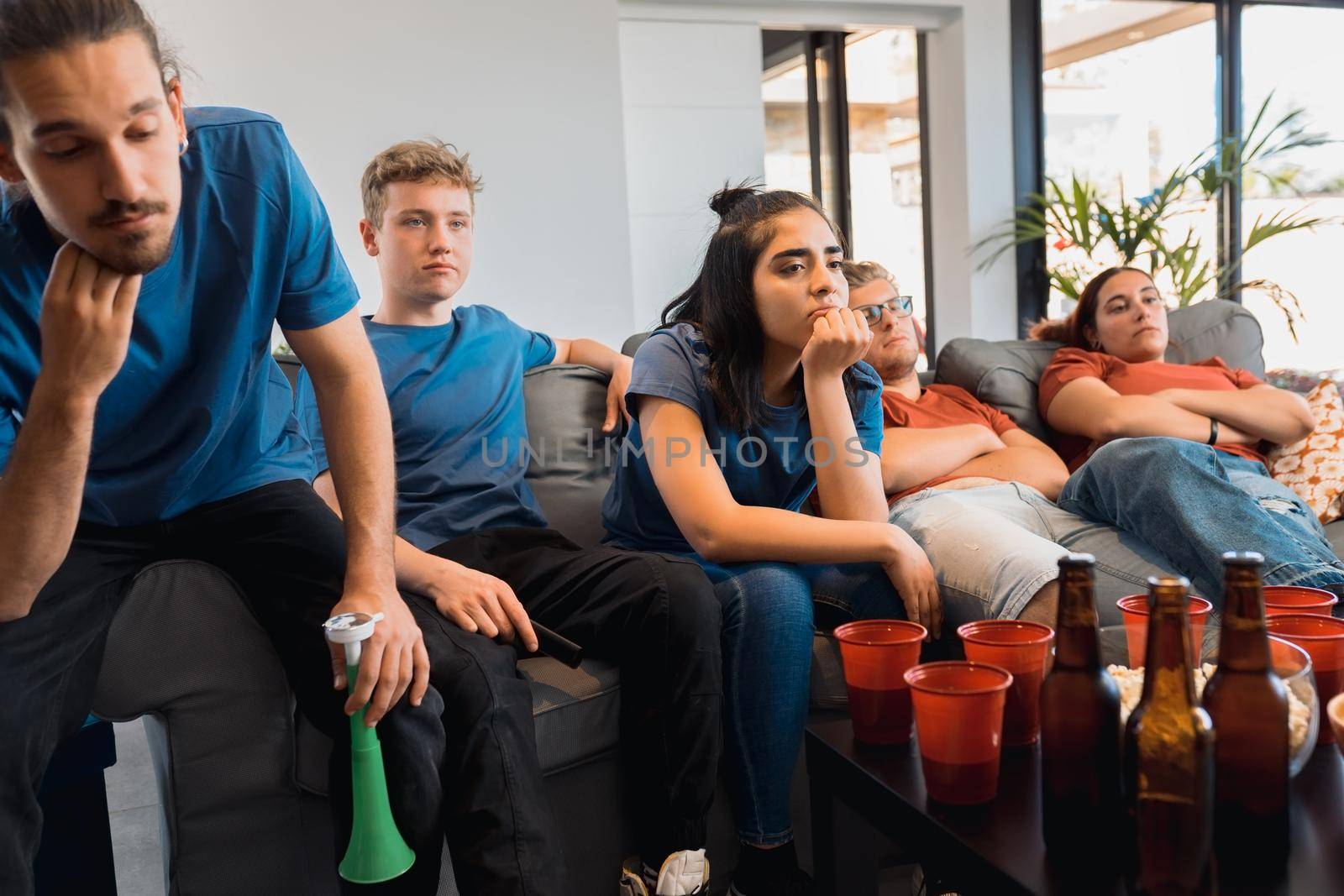 group of friends disappointed with their team, watching a football match on TV. bored young people at a party at home. by CatPhotography