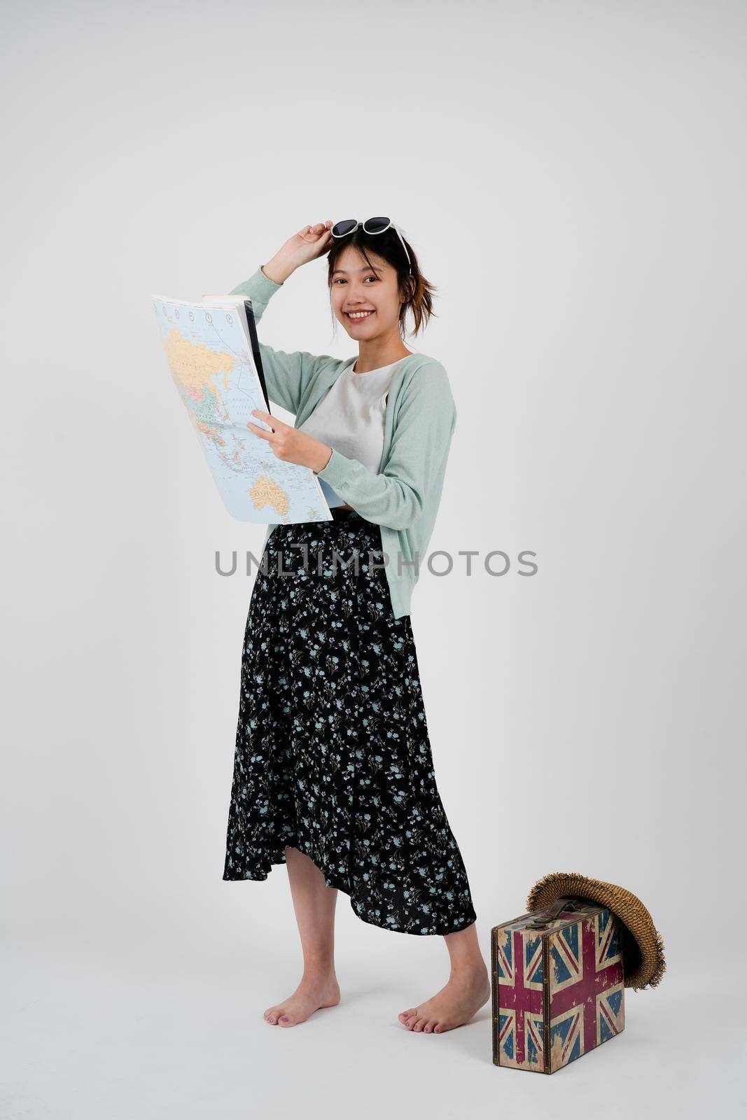 Positive smiling young asian female tourist searches for inspiring places, holds paper map, finds new sightseeing for discovering, wears round spectacles, isolated on white studio wall by nateemee