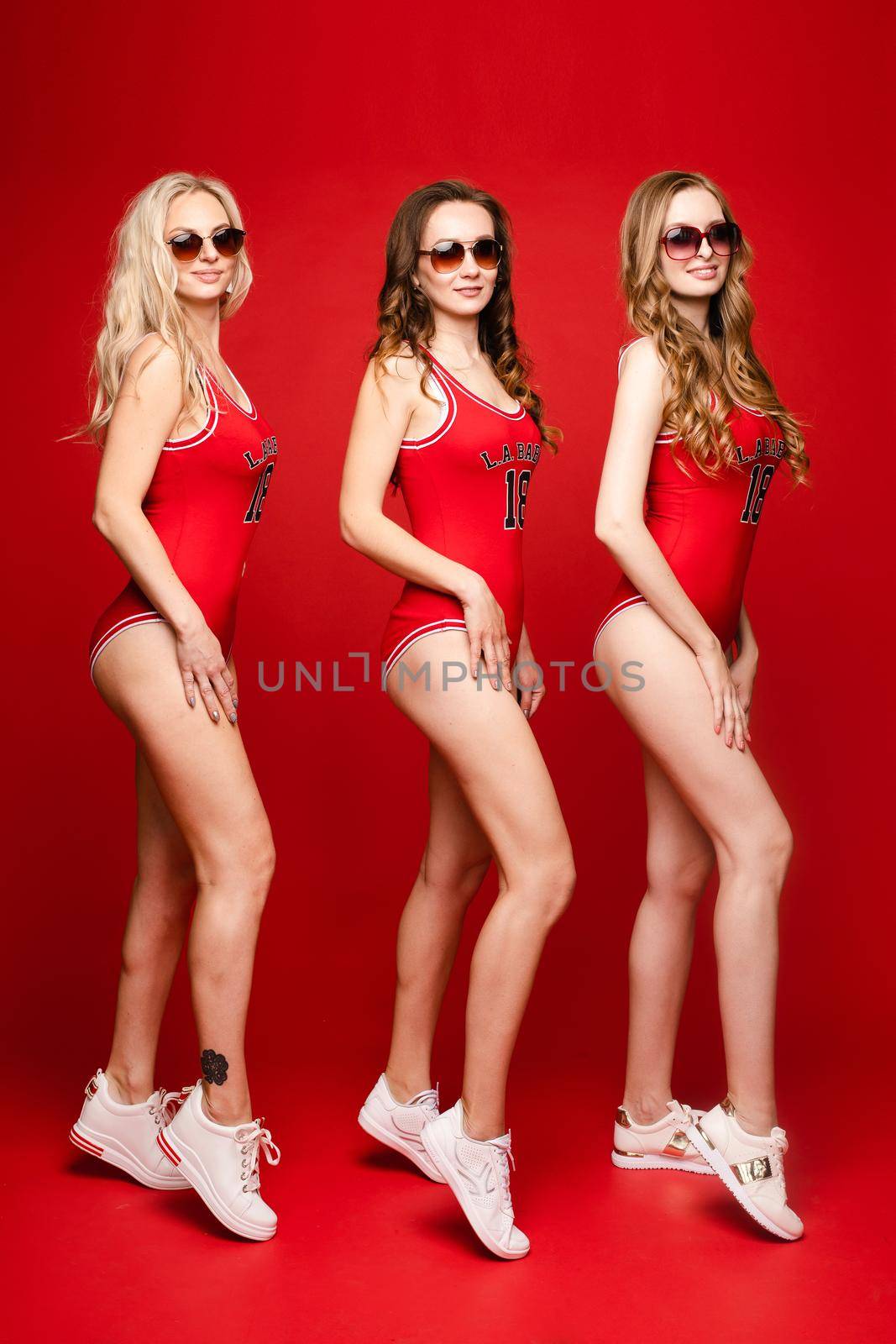 Three women in red swimsuits on a red background in the studio wearing sunglasses. by StudioLucky