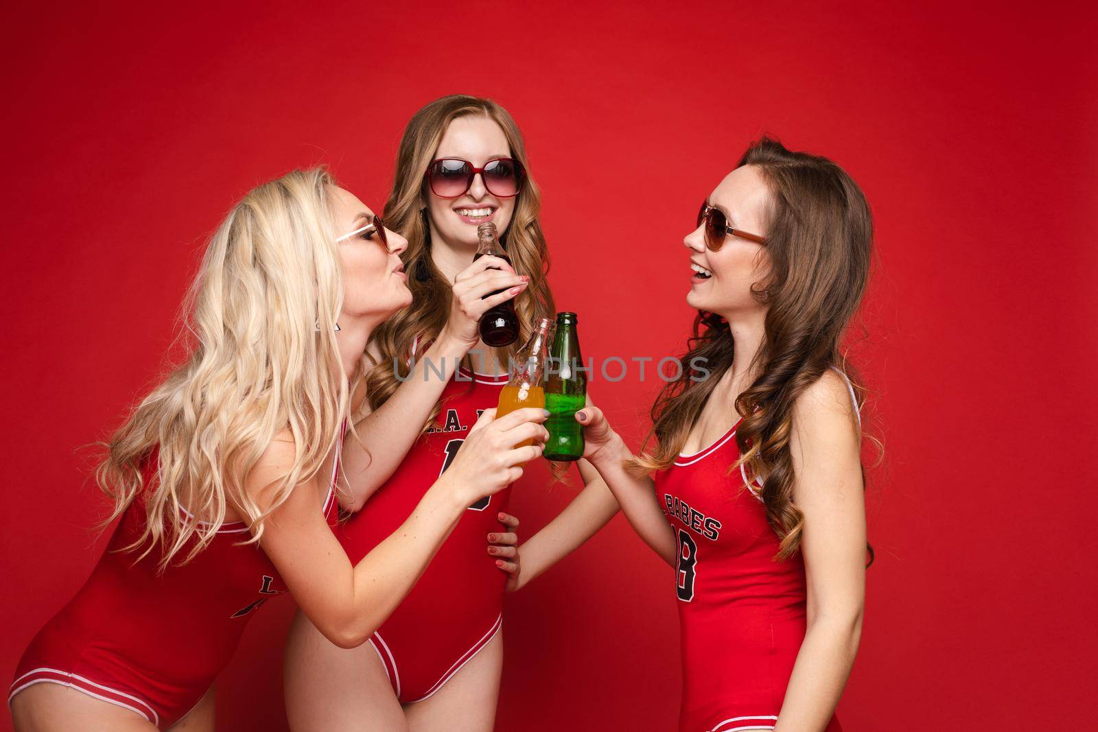 Beautiful and happy female friends in bright red swimsuits with chilled drinks in their hands on a red background, studio