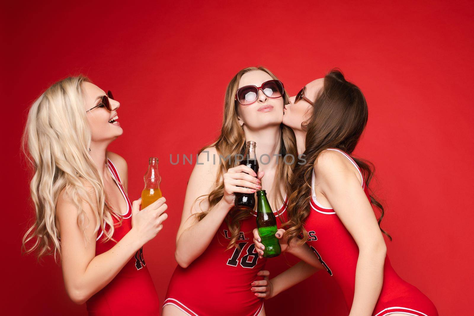 Beautiful and happy female friends in bright red swimsuits with chilled drinks in their hands on a red background, studio