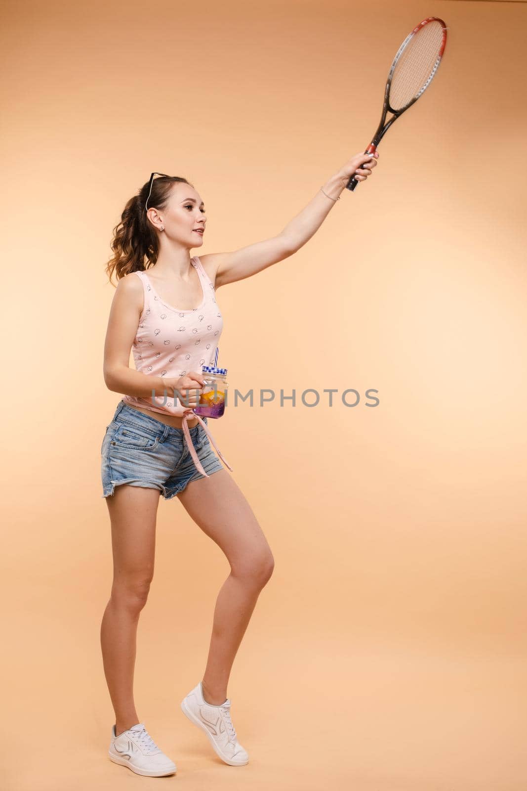 A beautiful woman in a bright outfit, denim shorts with a racket in her hands in the studio. Summer by StudioLucky