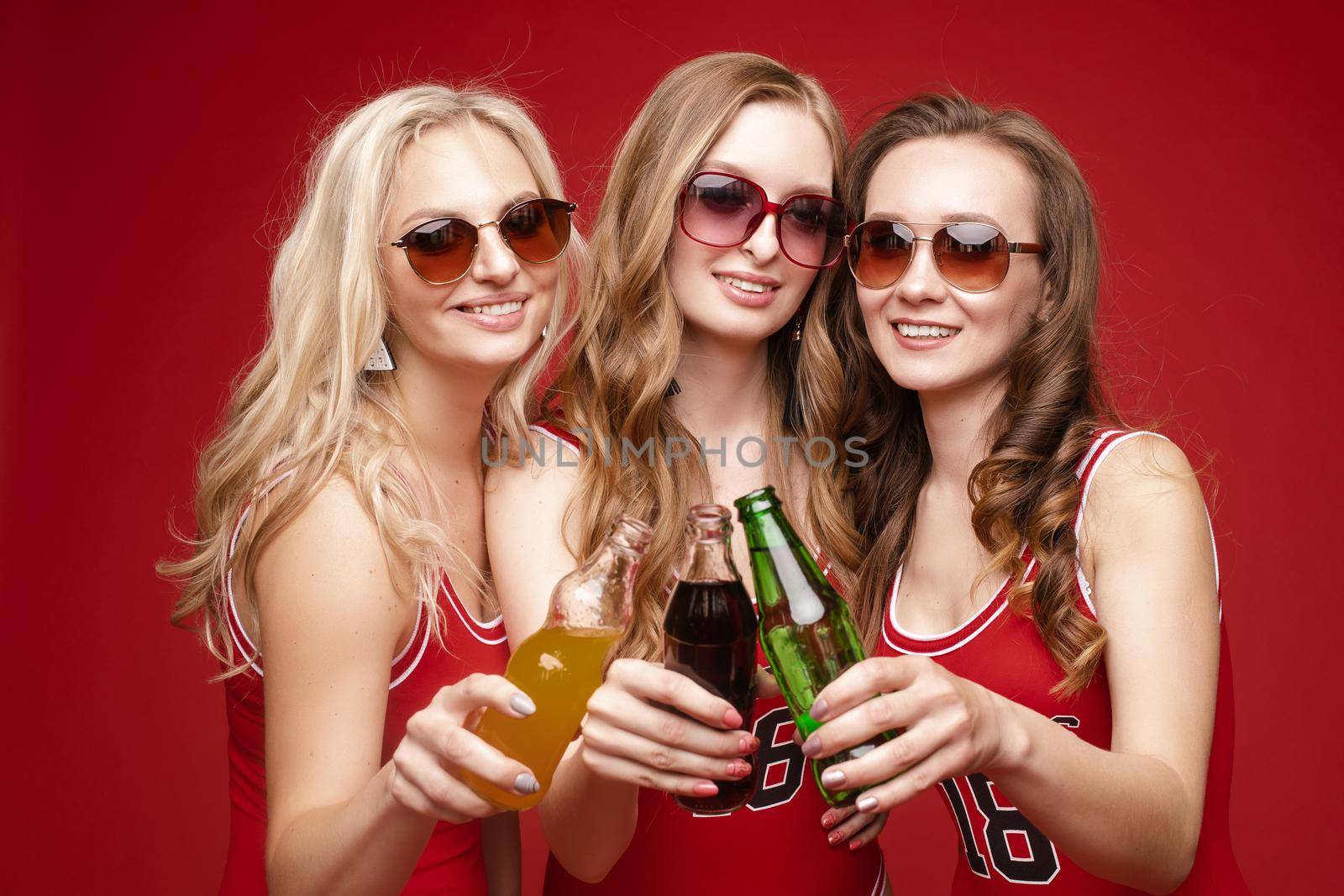 Beautiful and happy female friends in bright red swimsuits with chilled drinks in their hands on a red background, studio. by StudioLucky